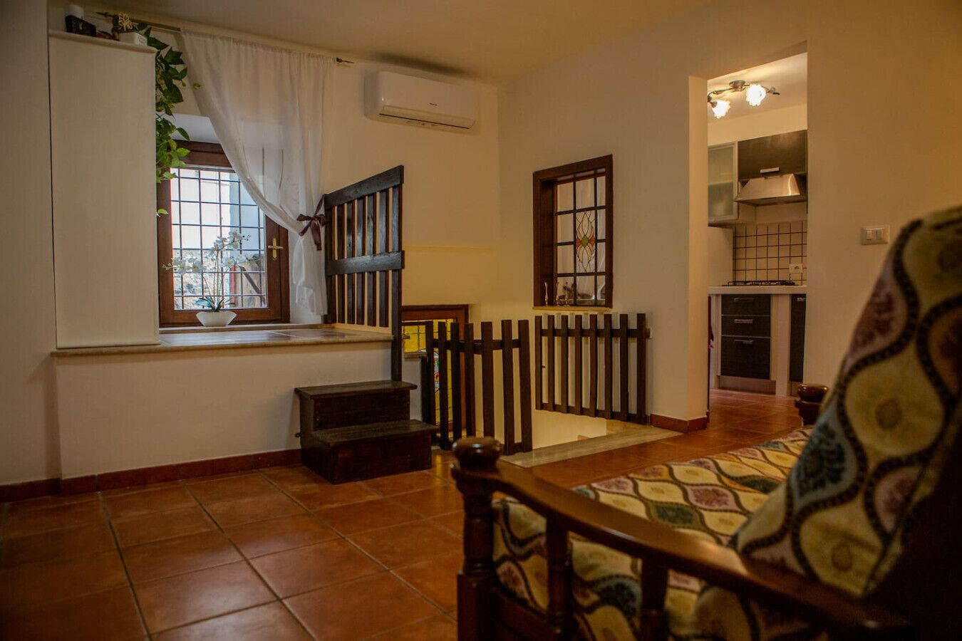 Un salon avec un banc en bois, une petite table, une fenêtre avec des rideaux, un escalier et un coin cuisine.