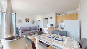 A modern living room with a sofa, dining table, and chairs. There is a kitchen area with cabinets and appliances. - Le Reflet Marin (Saint-Jean-de-Monts)