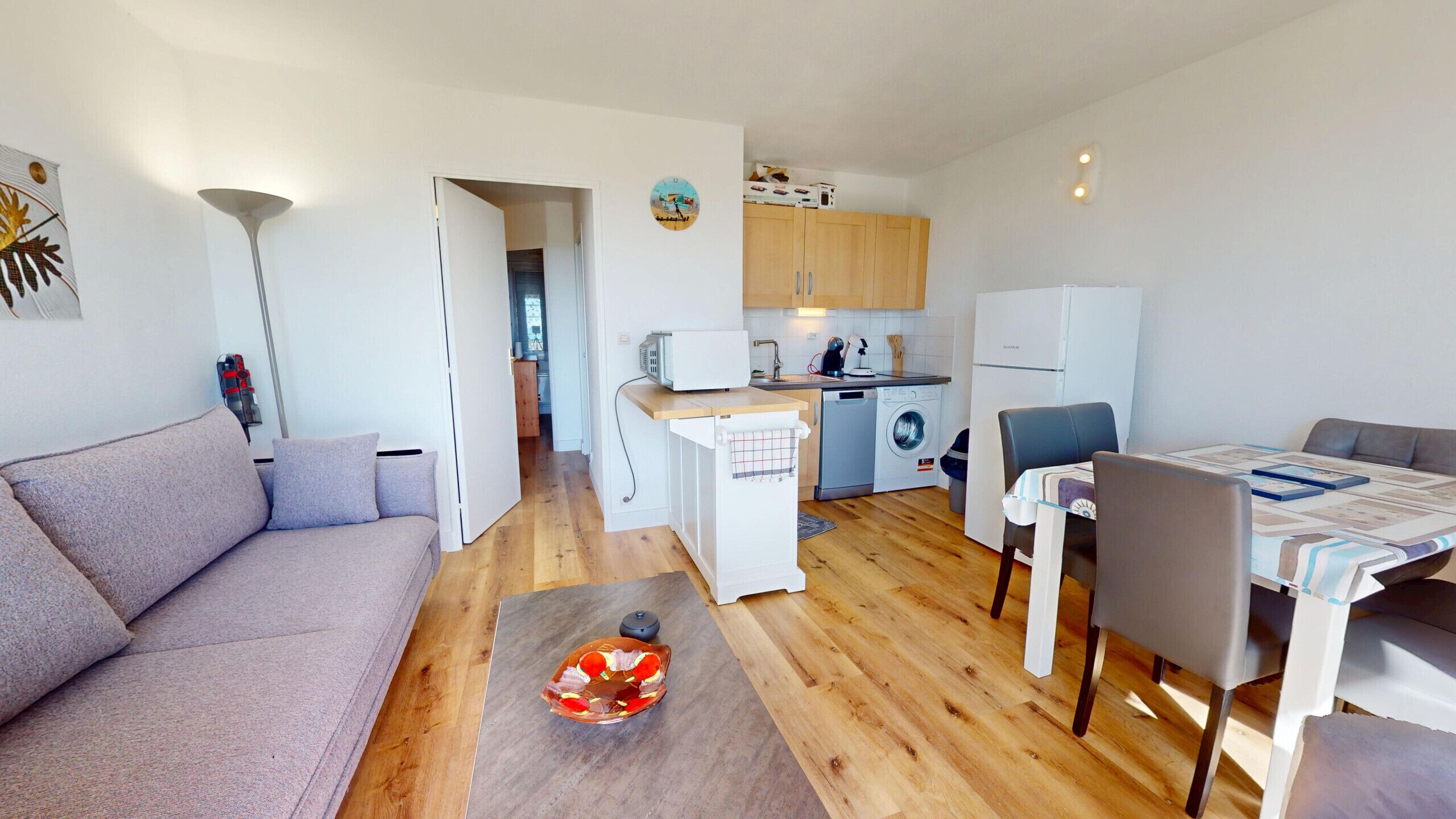 A compact living space with a sofa, dining table, chairs, a small kitchen area with a washing machine, and a refrigerator.