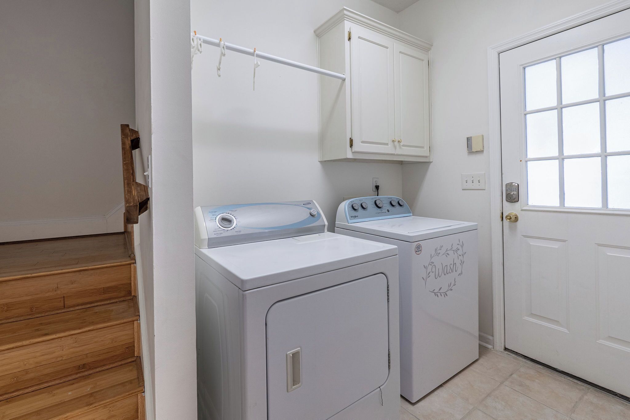 A laundry room with two white washing machines, a white cabinet above, and a white door with a window.