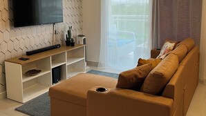 Ein modernes Wohnzimmer mit einem Flachbildfernseher an einer Wand mit sechseckigem Muster, einer Holzunterhaltungsecke, einem braunen Sofa mit Kissen und einem großen Fenster mit transparenten Vorhängen.