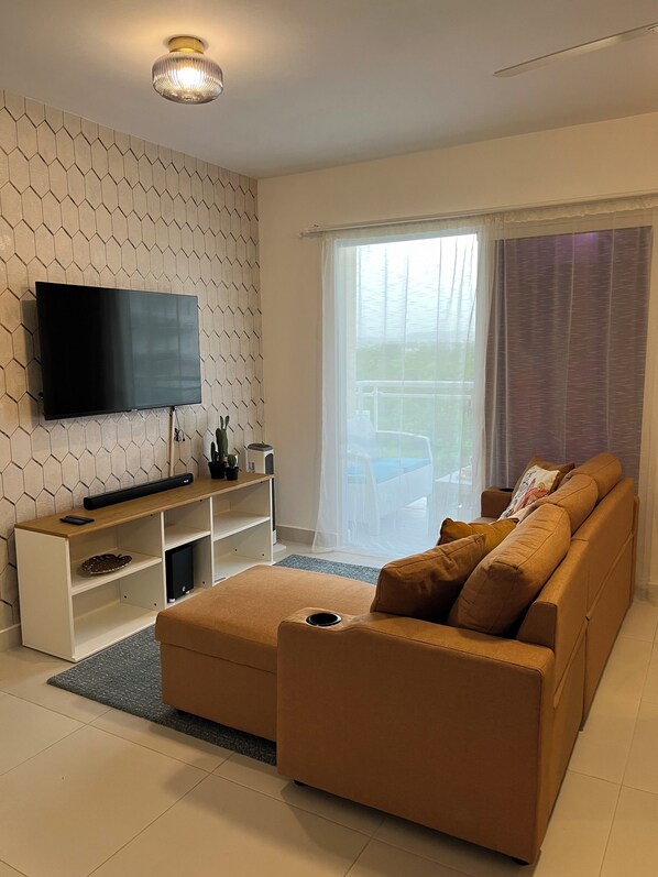A modern living room with a flat-screen TV mounted on a hexagonal-patterned wall, a wooden entertainment unit, a brown sofa with cushions, and a large window with sheer curtains.
