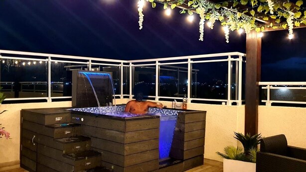 Un bain à remous extérieur agrémenté d’une cascade, entouré d’une pergola illuminée de guirlandes lumineuses, le tout offrant une vue sur le paysage urbain nocturne.