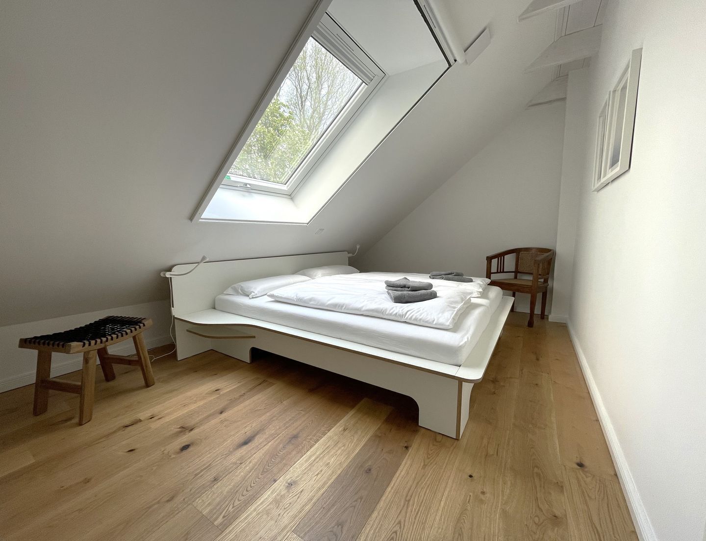 Habitación pequeña con techo blanco inclinado, una cama, una silla y un tragaluz.