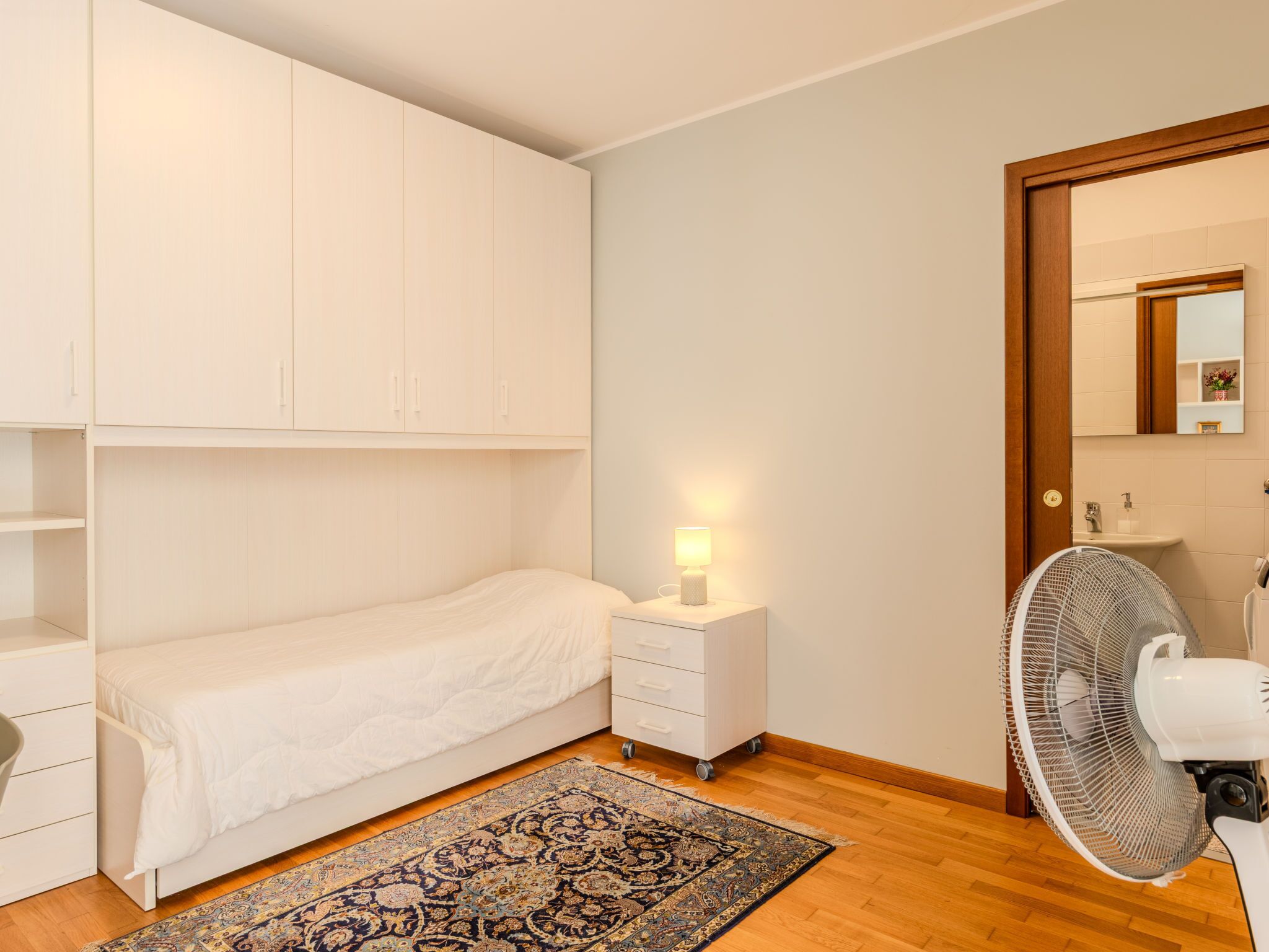 Et soveværelse med en seng, et natbord med en lampe, en ventilator og trægulv.