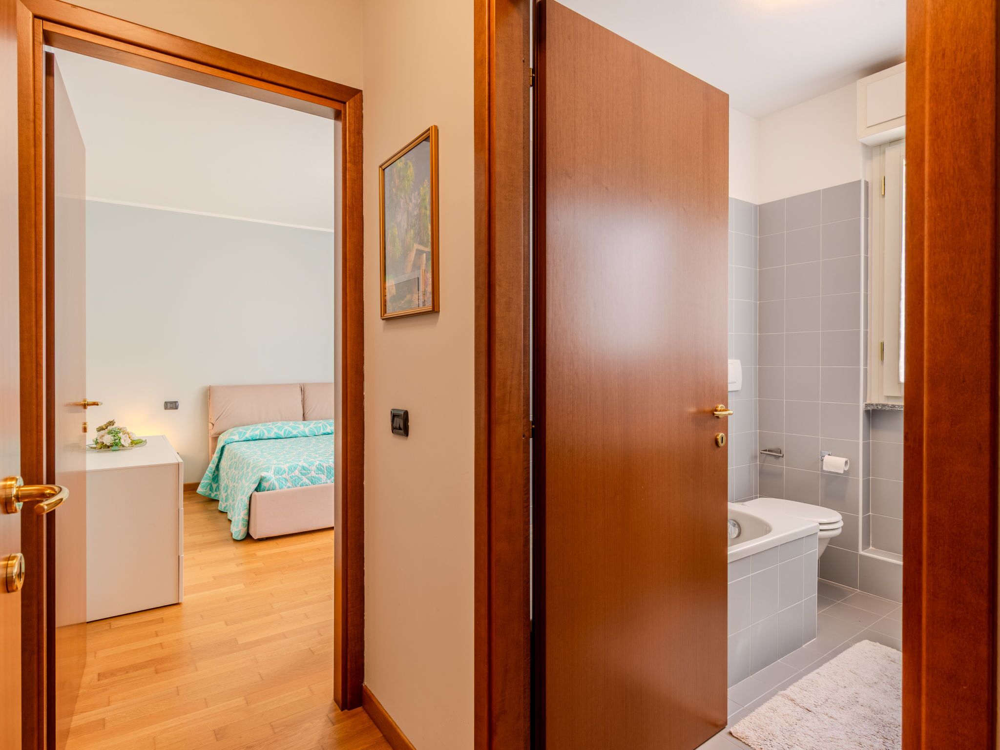Ett hotellrum med en dörr av trä, en säng med ett blått och vitt mönstrat överkast, ett sängbord med en blomsteruppsättning och ett badrum med ett vitt handfat och kaklade väggar.