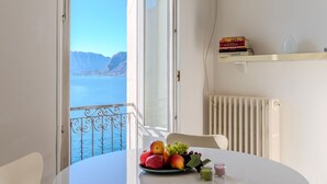 Una habitación con vista a un cuerpo de agua y montañas, una mesa con fruta, una silla y un radiador.