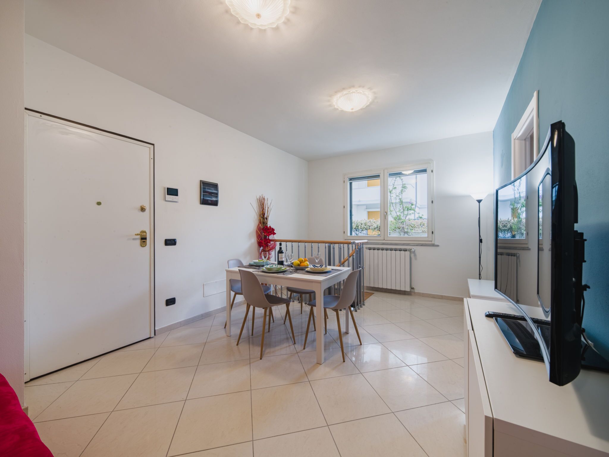A modern kitchen with white cabinets, a dining table with chairs, a TV, and a window with a view of greenery.
