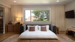 Ein Schlafzimmer mit einem großen Bett, einem Fenster mit Vorhängen, einem an der Wand befestigten Fernseher und einer Holzkommode.