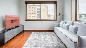 A modern living room with a grey sofa, a flat-screen TV, a wooden floor, and a large window with a city view. - GuestReady - Serene hideout in Leça da Palmeira (Porto)