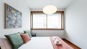 A bedroom with a large bed, a window with a view of buildings, a hanging light fixture, and a wall-mounted artwork. - GuestReady - Serene hideout in Leça da Palmeira (Porto)