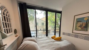 A bedroom with a bed, a window with curtains, a wall-mounted shelf with a decorative item, and a framed picture on the wall.