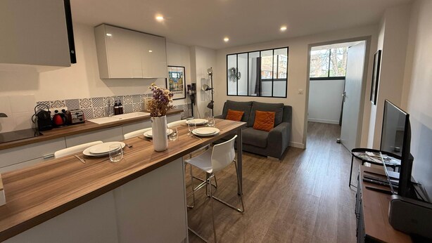 A modern kitchen with a wooden countertop, white cabinets, and a dining area with a grey sofa and orange cushions.