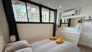 A hotel room with a bed, two pillows, a bedside table, a dresser, a mirror, and a window with a view of buildings.