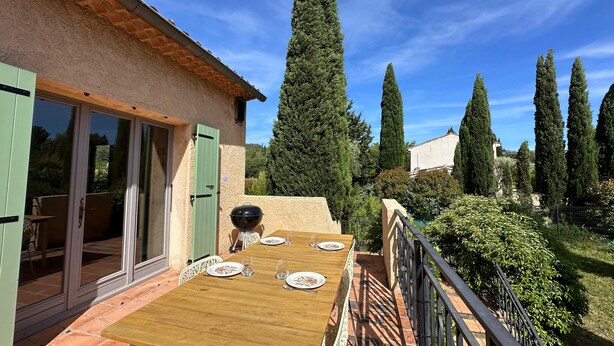 Una terrazza con un tavolo in legno apparecchiato per consumare un pasto, una porta scorrevole in vetro e vista su alti cipressi.