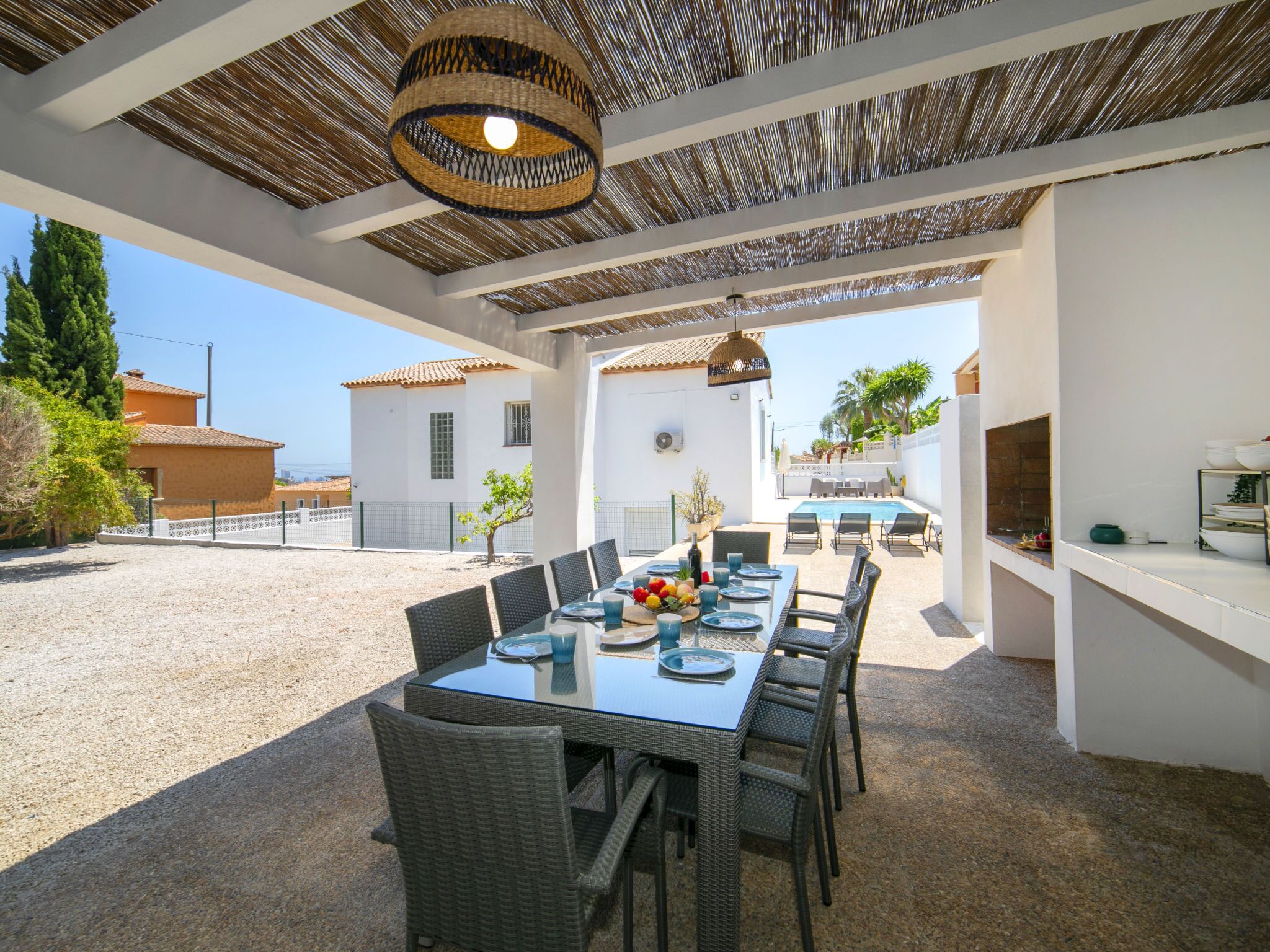 An outdoor dining area with a table set for six, surrounded by wicker chairs, under a woven ceiling.