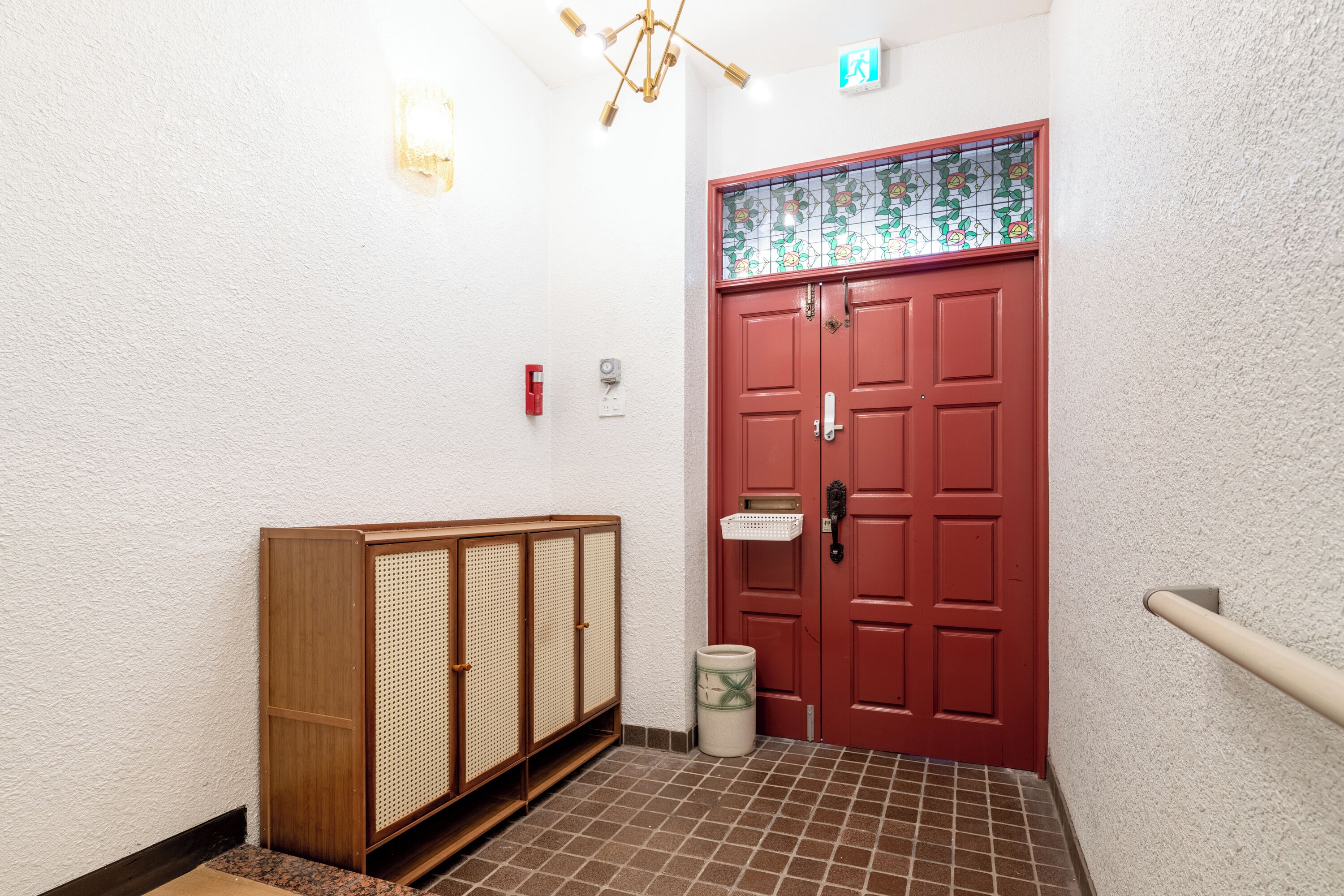 A hallway with a red door, a wooden cabinet, a trash can, and a tiled floor.