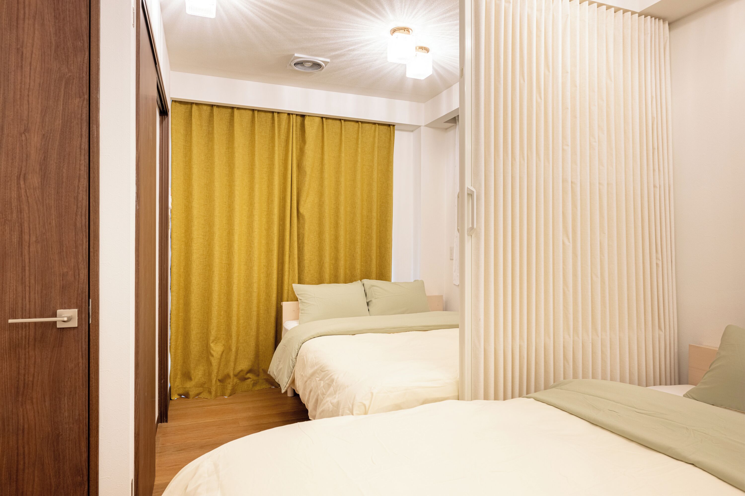 A bedroom with two beds, a wooden door, a yellow curtain, and a white ceiling with lights.