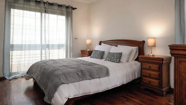 Una cama bien hecha con cabecera de madera, dos mesitas de noche con lámparas y un ventanal con cortinas de gasa.