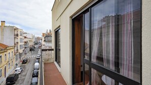 Un balcón estrecho con vistas a una calle flanqueada por edificios, un coche aparcado y un cielo despejado.