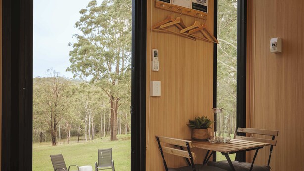 A room with a wooden table and chairs, a hanging clothes rack, and a view of a grassy area with trees.