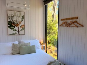 Een slaapkamer met een bed, kussens, een wandairconditioner, een raam met uitzicht op groen, en houten kledinghangers aan de muur.