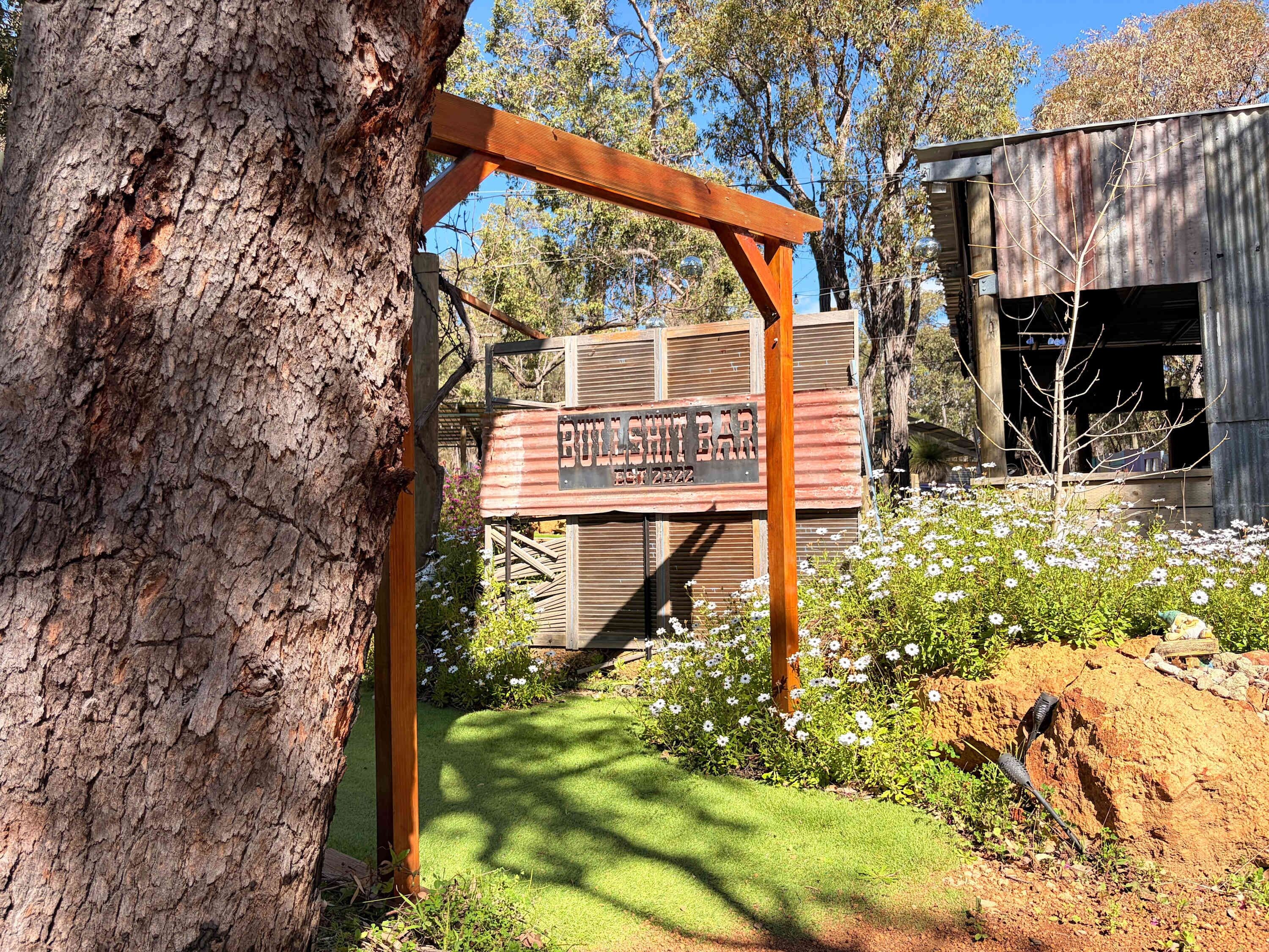 Un letrero de madera rústico que dice 'BULLSHIT BAR', con una reja metálica y un árbol en primer plano.
