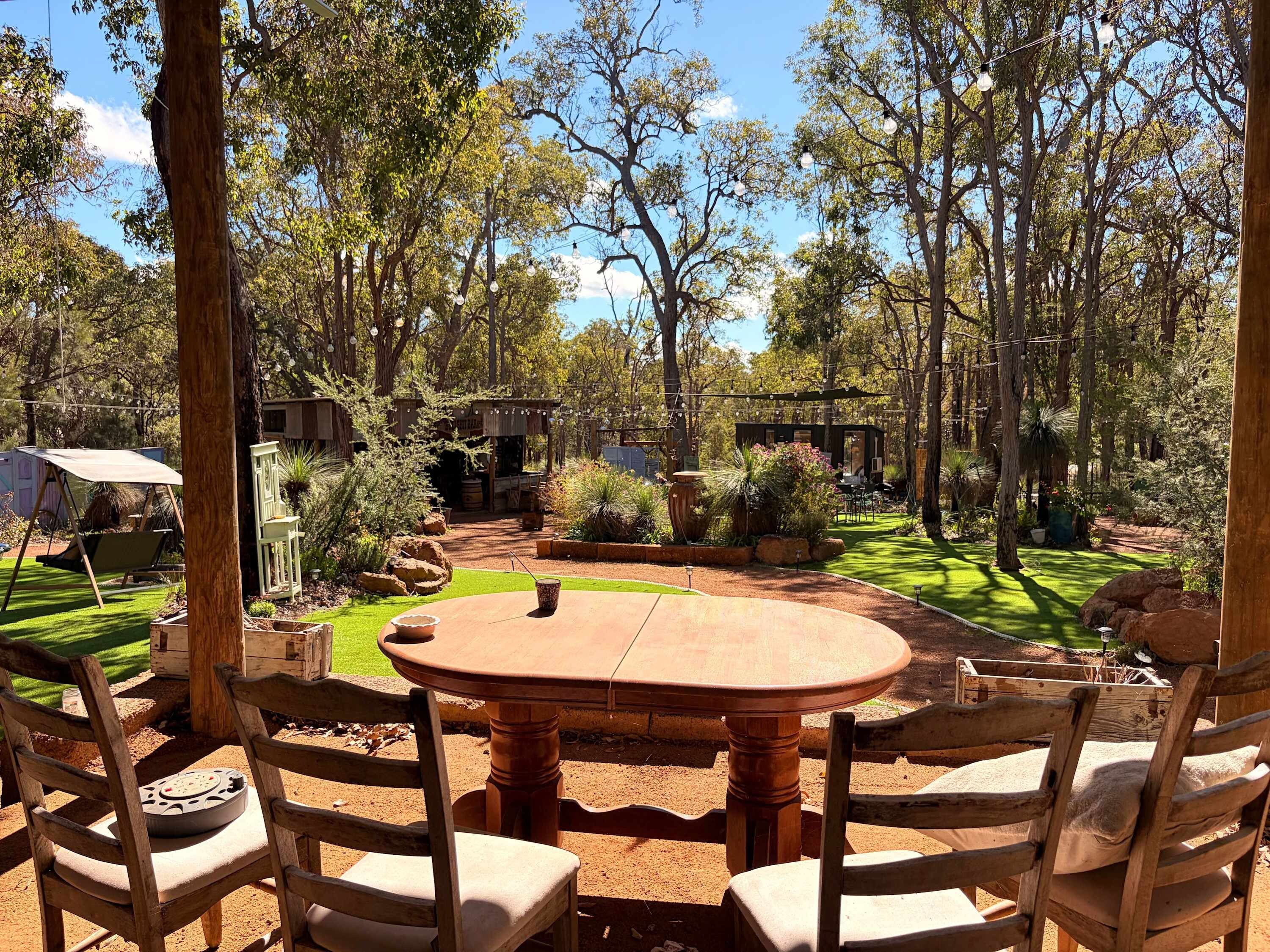 Een terras buiten met een ronde houten tafel en stoelen, omgeven door bomen en een verzorgd gazon.