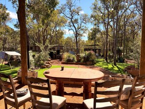 Een terras buiten met een ronde houten tafel en stoelen, omgeven door bomen en een verzorgd gazon.