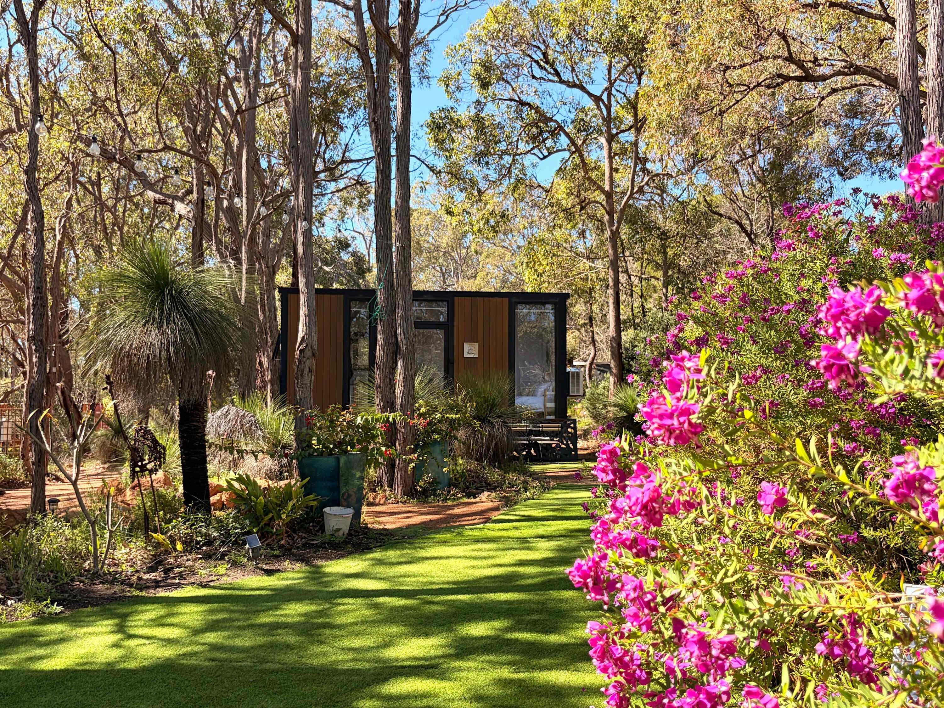 A garden with a pathway leading to a wooden structure, surrounded by trees and flowering plants.