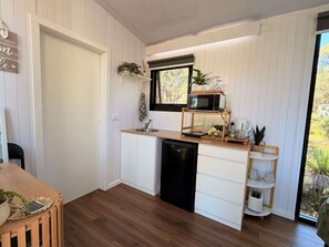 Een compacte keuken met een ingebouwde vaatwasser, een magnetron op een houten plank, en een raam met uitzicht op groen.