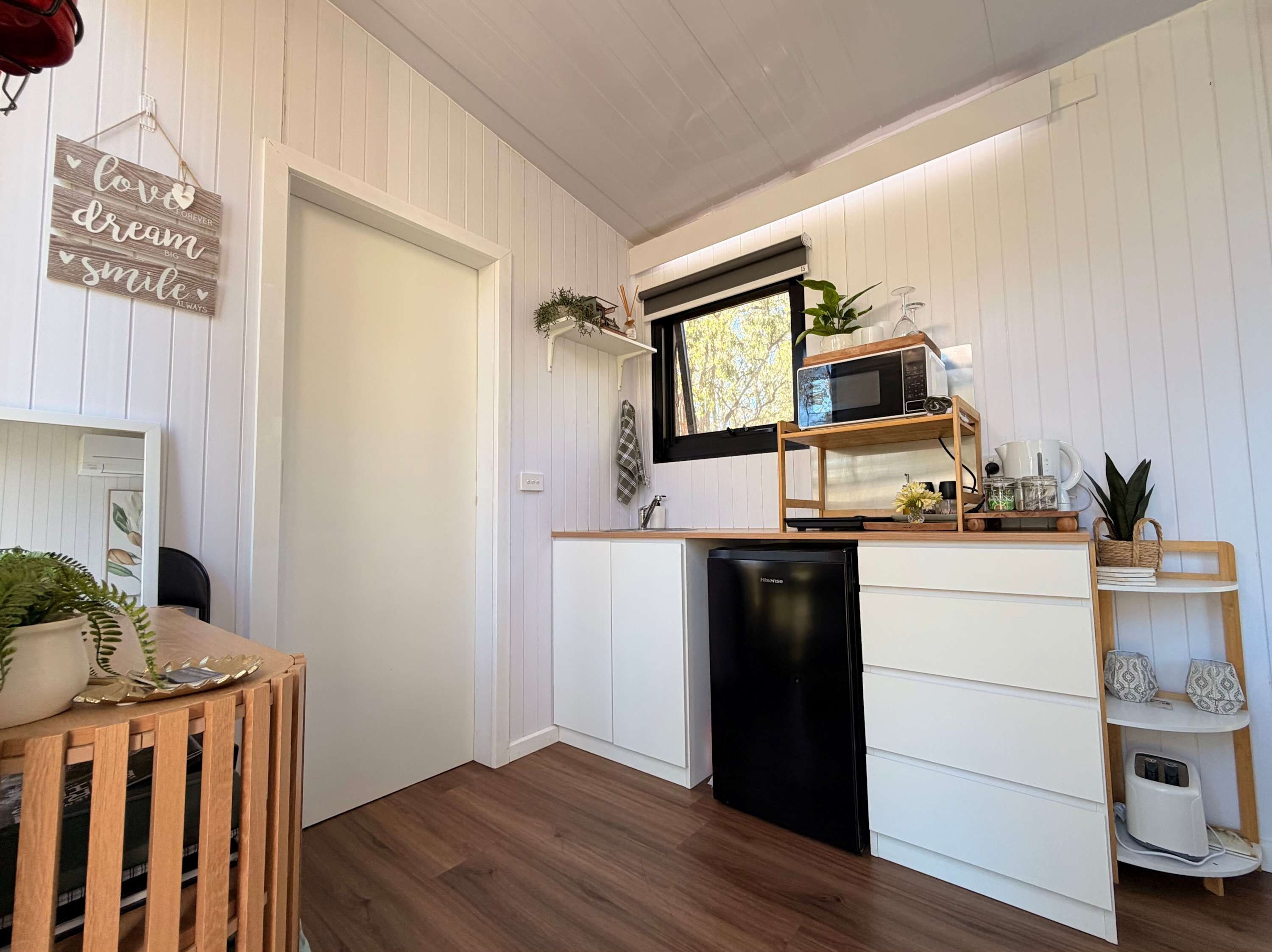 Een compacte keuken met een zwarte vaatwasser, een magnetron en een houten plank met diverse spullen.