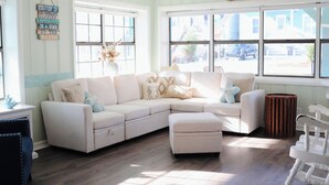 A cozy living room with a sectional sofa, a small ottoman, and a rocking chair. There are large windows with a view of the street outside. - Cozy Retreat , steps to the beach, heated pool , gulf views & huge party Deck (Panama City Beach)