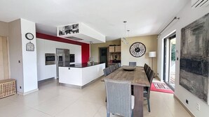 Une cuisine moderne avec des meubles blancs, un mur d’accent rouge et un coin repas comprenant une grande table et des chaises en bois.