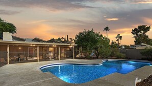 Una piscina con bordo curvo, circondata da un solarium in cemento. C’è un’area patio coperta con sedie e un tavolo, e una palma alta sullo sfondo.