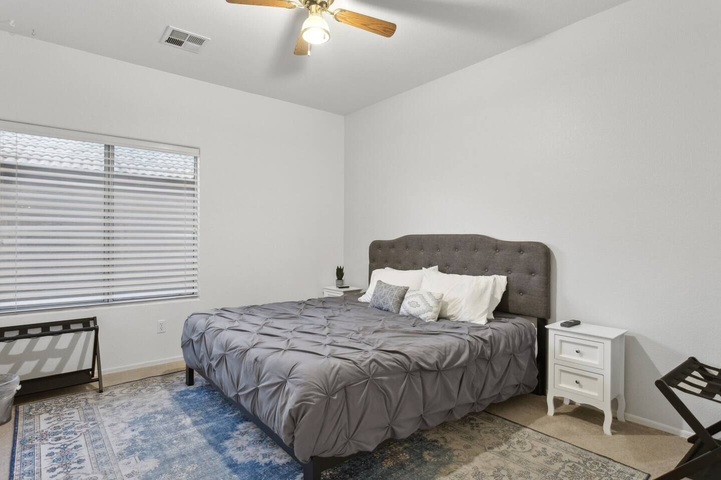 Une chambre à coucher comprenant un lit, une table de chevet, une chaise, un ventilateur de plafond et une fenêtre avec des stores.