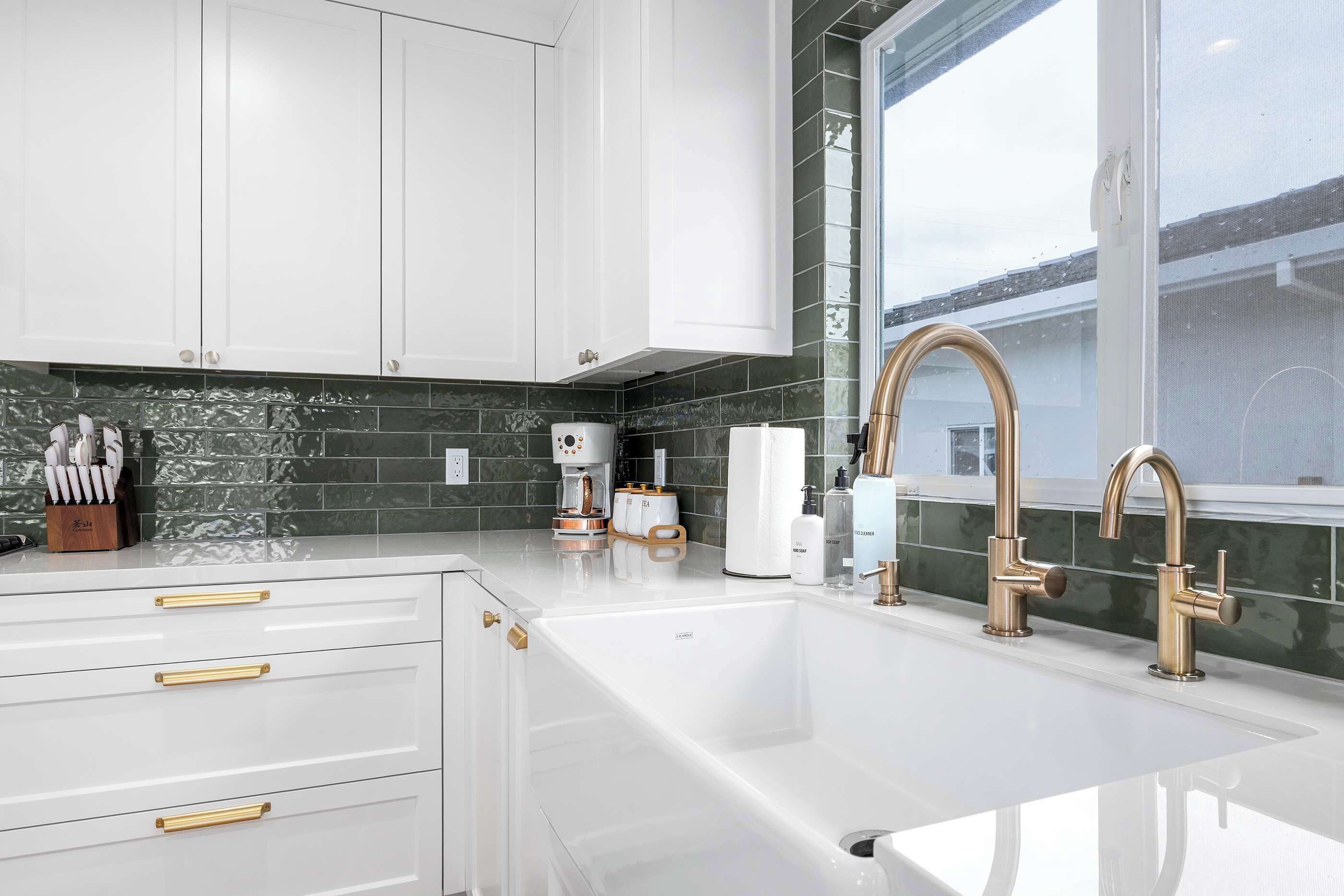 Une cuisine moderne avec des meubles blancs, un évier en acier inoxydable et une fenêtre donnant sur un immeuble.