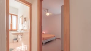 A narrow hallway leading to a bedroom with a bed, a ceiling fan, and a bathroom with a sink and mirror. - APPARTAMENTI GLI ORIZZONTI - ORIZZONTI N QUADRI 1 (Marina di Campo)