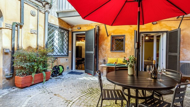 Eine Terrasse im Freien mit einem runden Tisch, Stühlen, einem roten Sonnenschirm und einem Blumentopf.