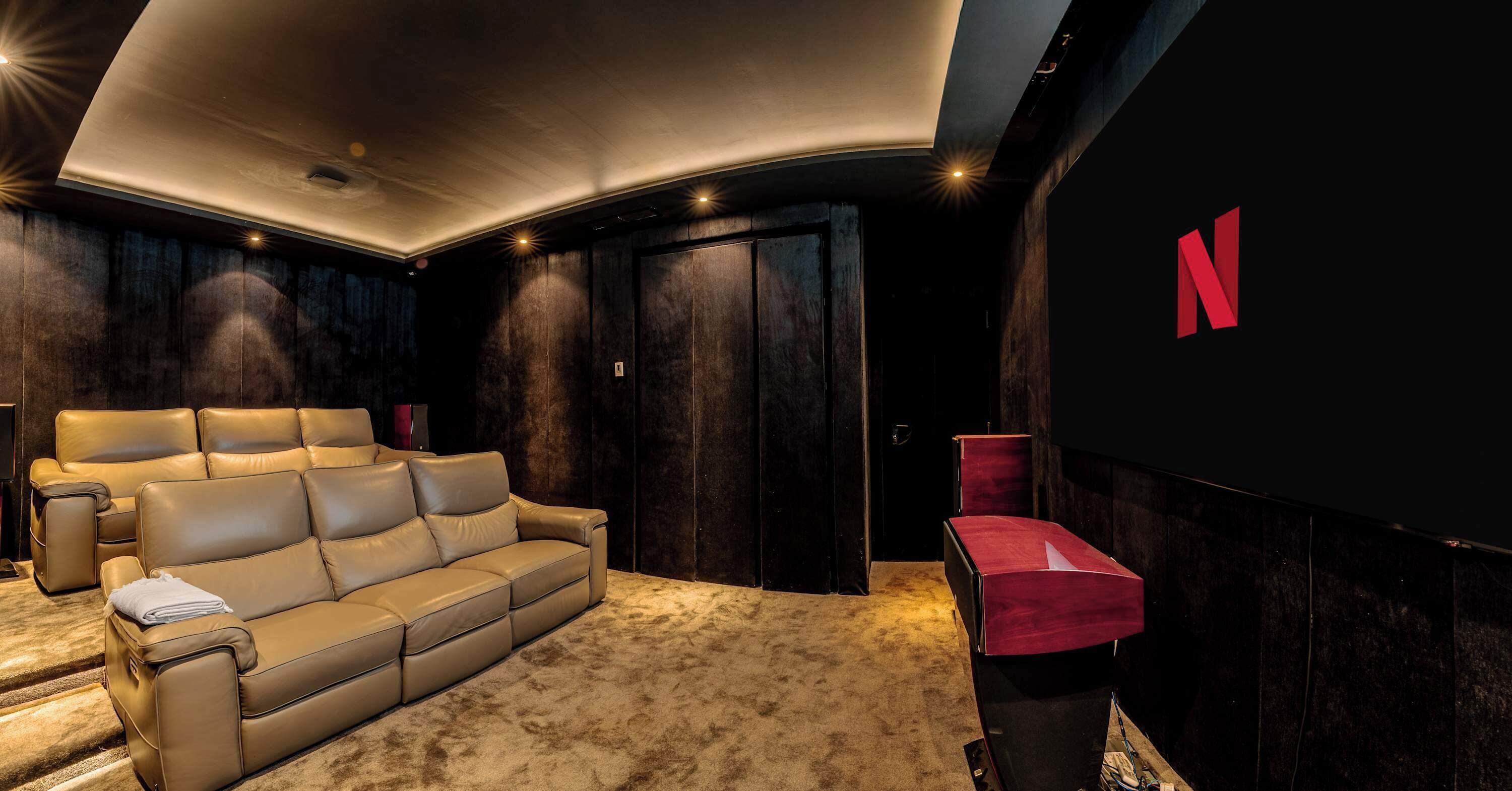Une salle de cinéma maison avec un grand canapé en cuir, un haut-parleur rouge et un écran mural affichant le logo de Netflix.