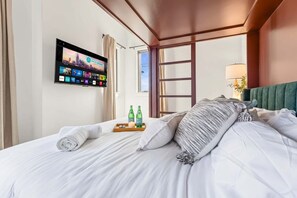 Ein Hotelzimmer mit Bett, Flachbildfernseher an der Wand, Fenster und Nachttisch mit Flaschen und Tablett.