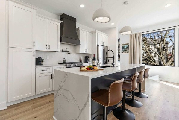 A modern kitchen with white cabinets, a central island with bar stools, and a large window offering a view of trees.