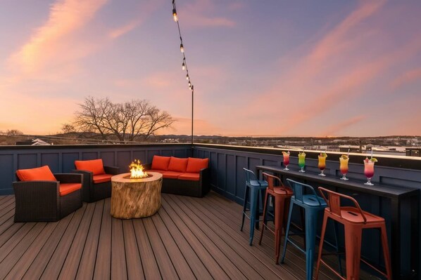 A rooftop deck with outdoor seating, a fire pit, and a bar area with colorful drinks.
