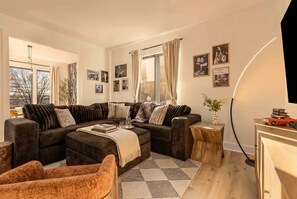 Ein modernes Wohnzimmer mit einer Eckcouch, einem Couchtisch und einem Holznachttisch. An der Wand befinden sich gerahmte Bilder und auf dem Nachttisch steht eine Pflanze.