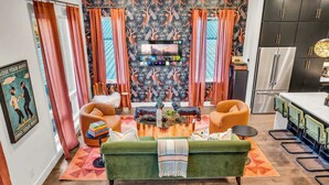 A cozy living room with a green sofa, orange armchairs, and a coffee table. A flat-screen TV is mounted on the wall, and there is a kitchen area with a refrigerator and cabinets.