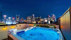 A rooftop hot tub with a city skyline view, wooden deck, and ambient lighting.