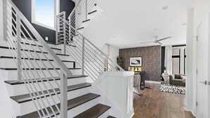 Un escalier moderne avec des rampes blanches et des marches en bois foncé mène à un espace de vie doté d’une cheminée, d’un ventilateur de plafond et d’une enseigne indiquant « BIBLIOTHÈQUE ».