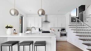 Une cuisine moderne avec des meubles blancs, un îlot central doté de tabourets de bar, des suspensions en acier inoxydable et un escalier avec des rampes en verre.
