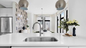 Une cuisine moderne dotée d’un réfrigérateur en acier inoxydable, de meubles blancs, d’un plan de travail blanc, d’un évier à deux bacs et de luminaires suspendus.