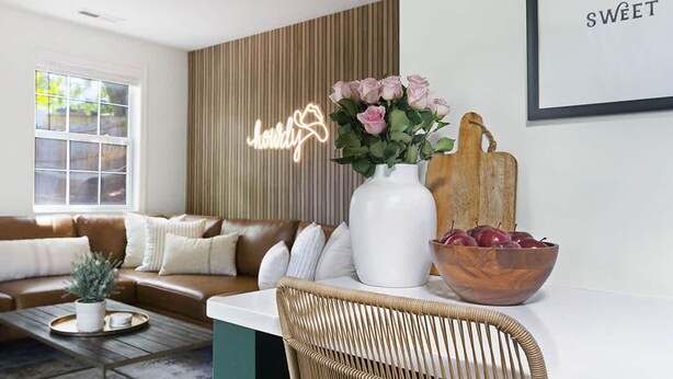A modern living room with a brown leather sofa, a white coffee table with a tray, a wicker chair, a white vase with flowers, and a neon sign on the wall. - Harmonic & Songbird Bungalows - 2, 2 bedrooms! (Nashville)